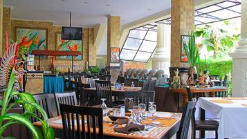 A restaurant with multiple tables and chairs, a television mounted on the wall, and tropical decor.