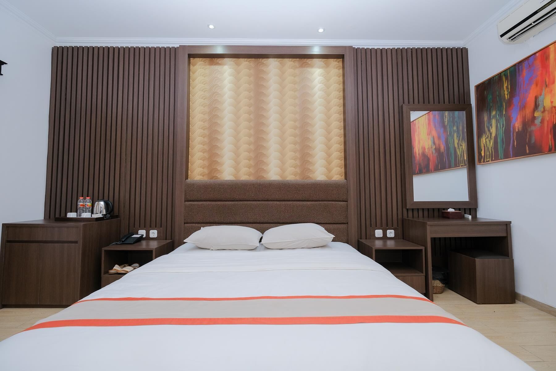 Una camera d'albergo con un letto grande, una testiera in legno, due comodini, un tavolino da notte, un'ampia finestra con tende e un quadro astratto dai colori vivaci appeso al muro.