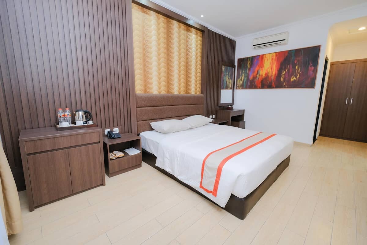 Een hotelkamer met een groot bed, een houten hoofdeinde, een nachtkastje met een telefoon en waterflessen, een bureau met een stoel, en een schilderij aan de muur.