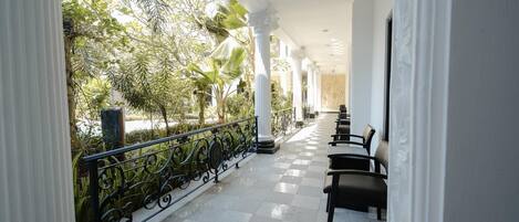 A covered outdoor area with a checkered floor, white columns, and a railing with greenery in the background.
