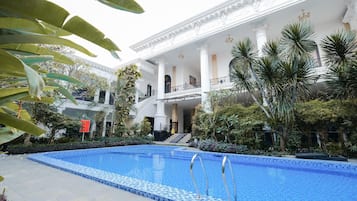 A pool area with a clear blue pool, surrounded by lush greenery and a grand building with columns and balconies.