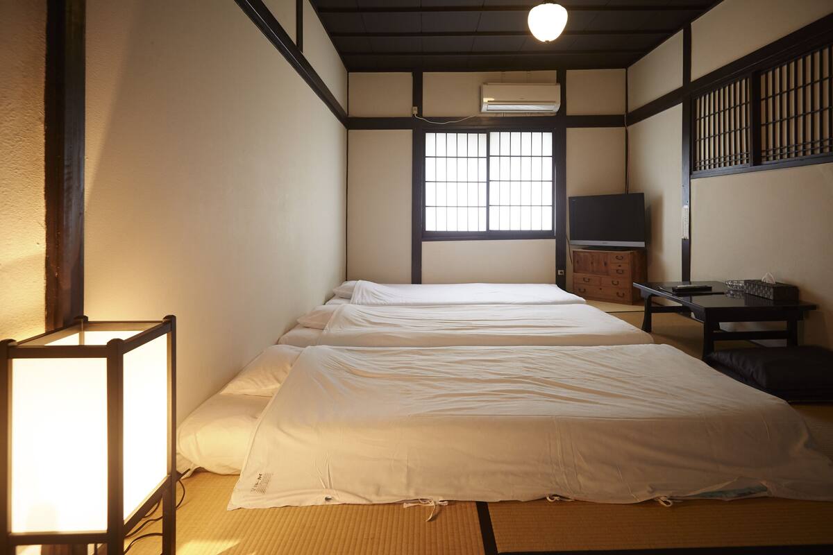 Quarto japonês tradicional com tatames, futon, mesa baixa e uma cadeira, e porta deslizante.
