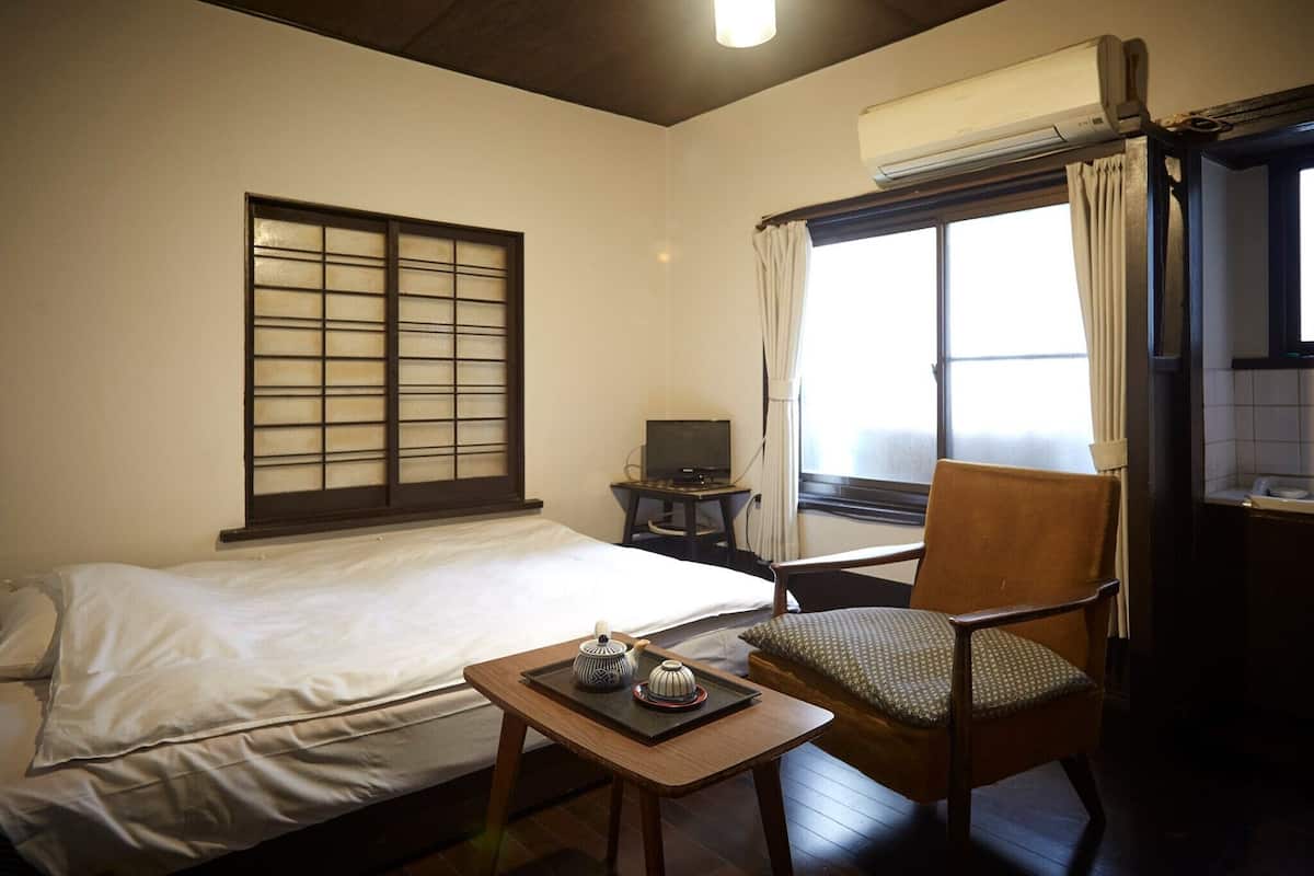 Quarto de hotel com uma cama, uma cadeira, uma mesinha com jogos de chá, uma televisão em uma mesinha lateral e uma janela com cortinas.