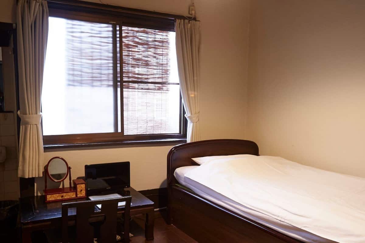 Um quarto pequeno e bem arrumado com uma cama de solteiro, uma escrivaninha com cadeira, uma mesinha de cabeceira com espelho e uma janela com cortinas.