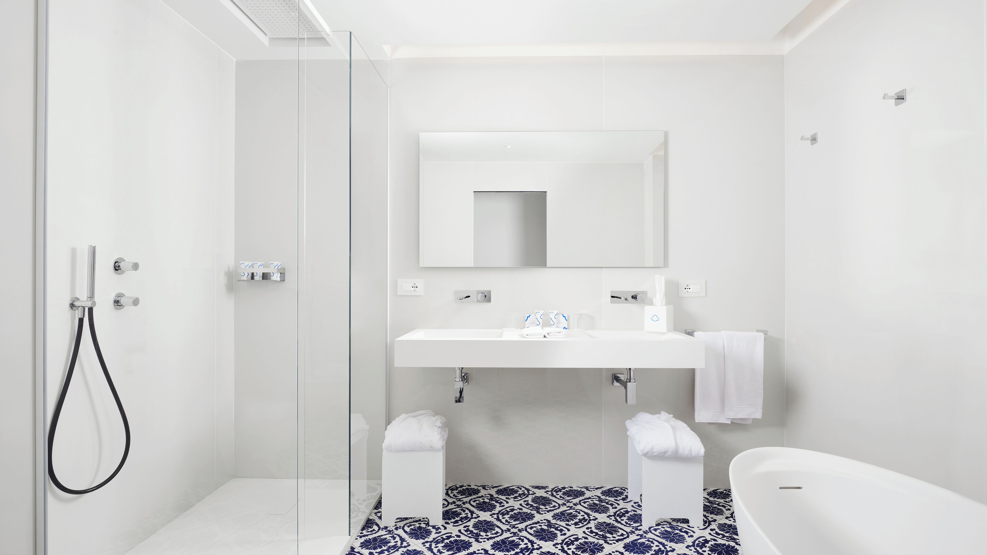 A modern bathroom with a glass shower enclosure, a white sink with a mirror above, and a bathtub with a blue and white patterned floor.