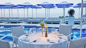 A poolside dining area with round tables set for two, blue umbrellas, and lounge chairs in the background.