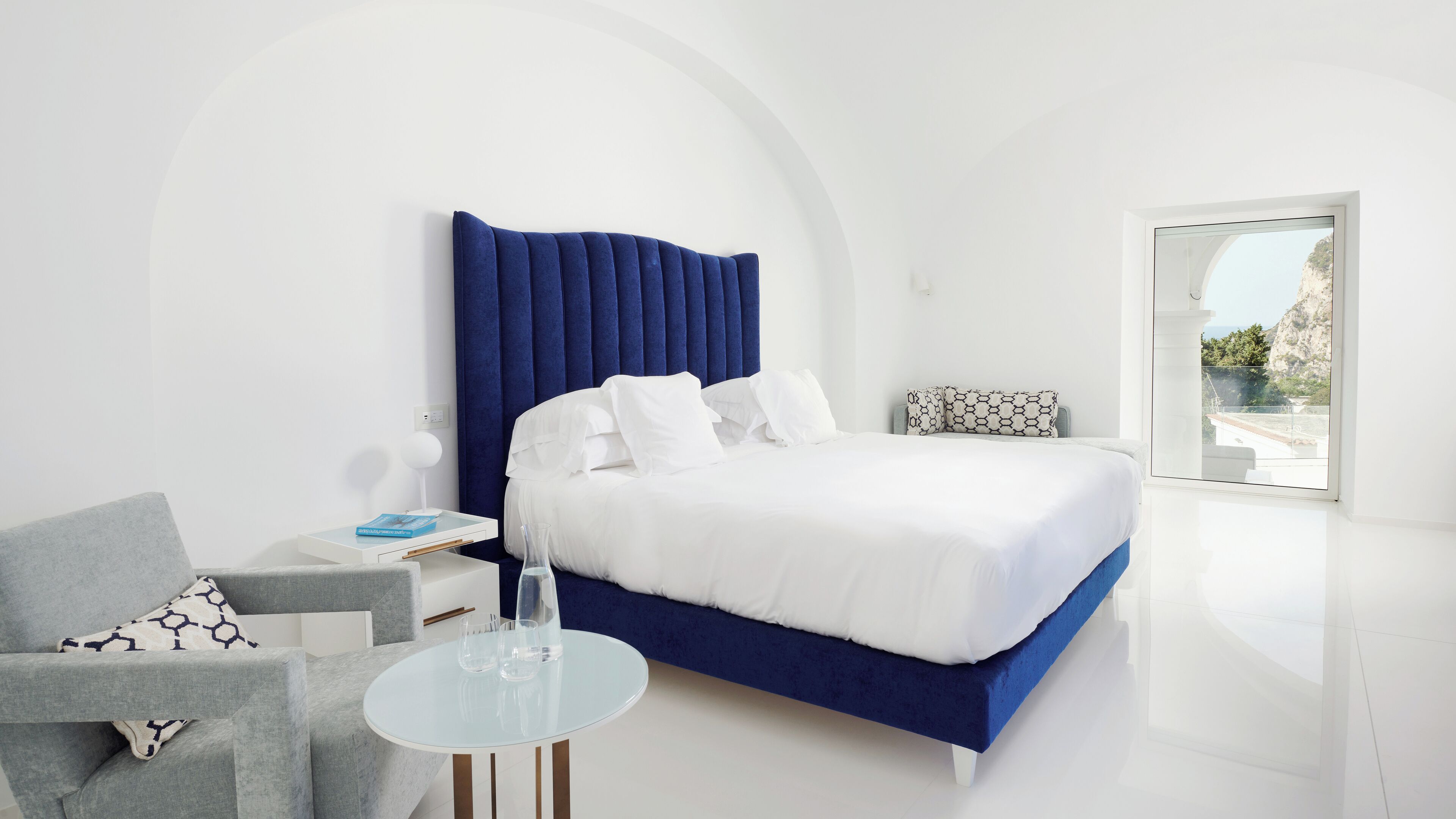 A modern bedroom with a large bed, a glass table, and a grey armchair.