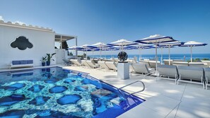 A rooftop pool with clear blue water, lounge chairs, umbrellas, and a view of the ocean.