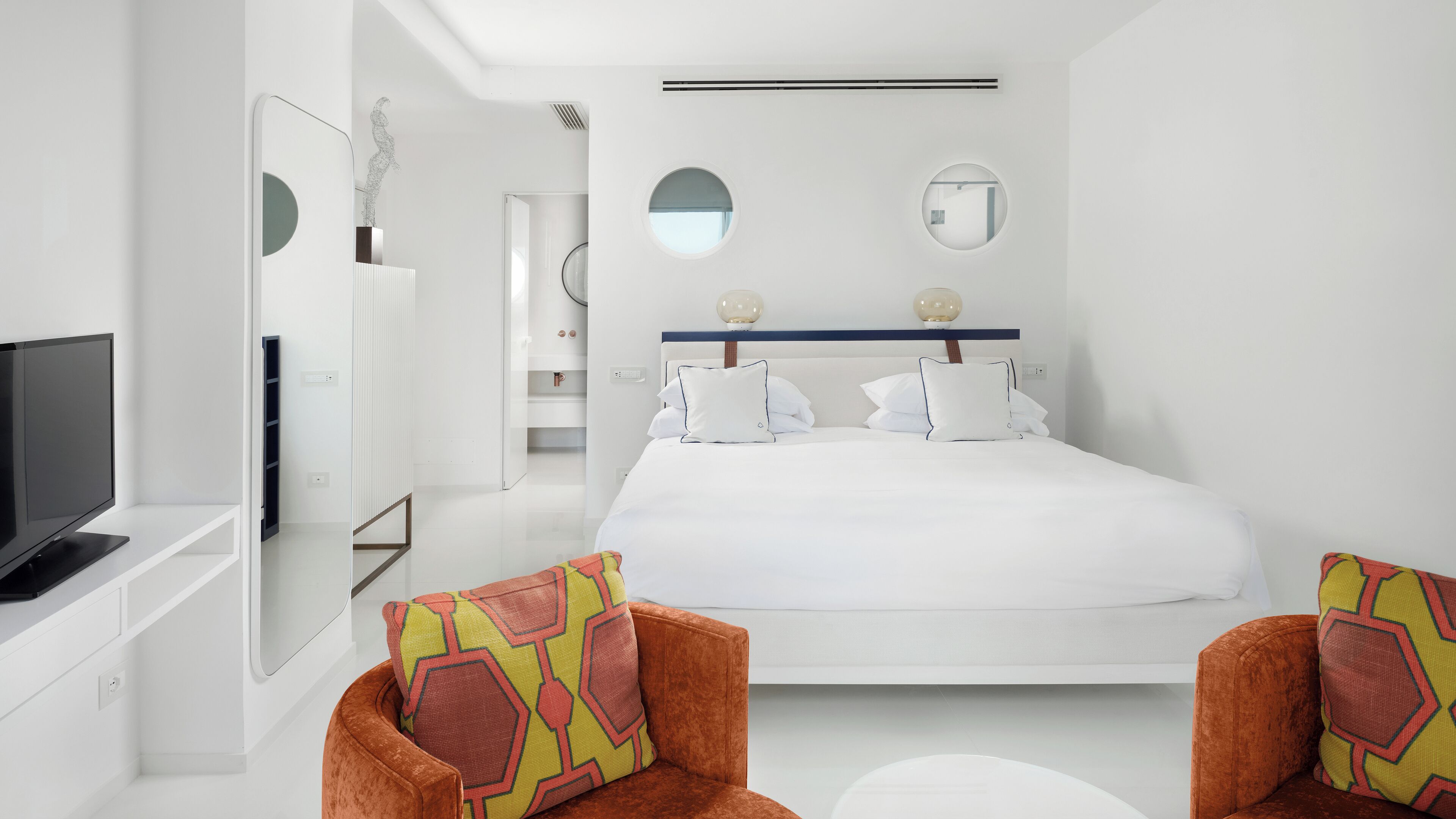 A modern hotel room with a large bed, two armchairs, a TV, and a mirror on the wall.