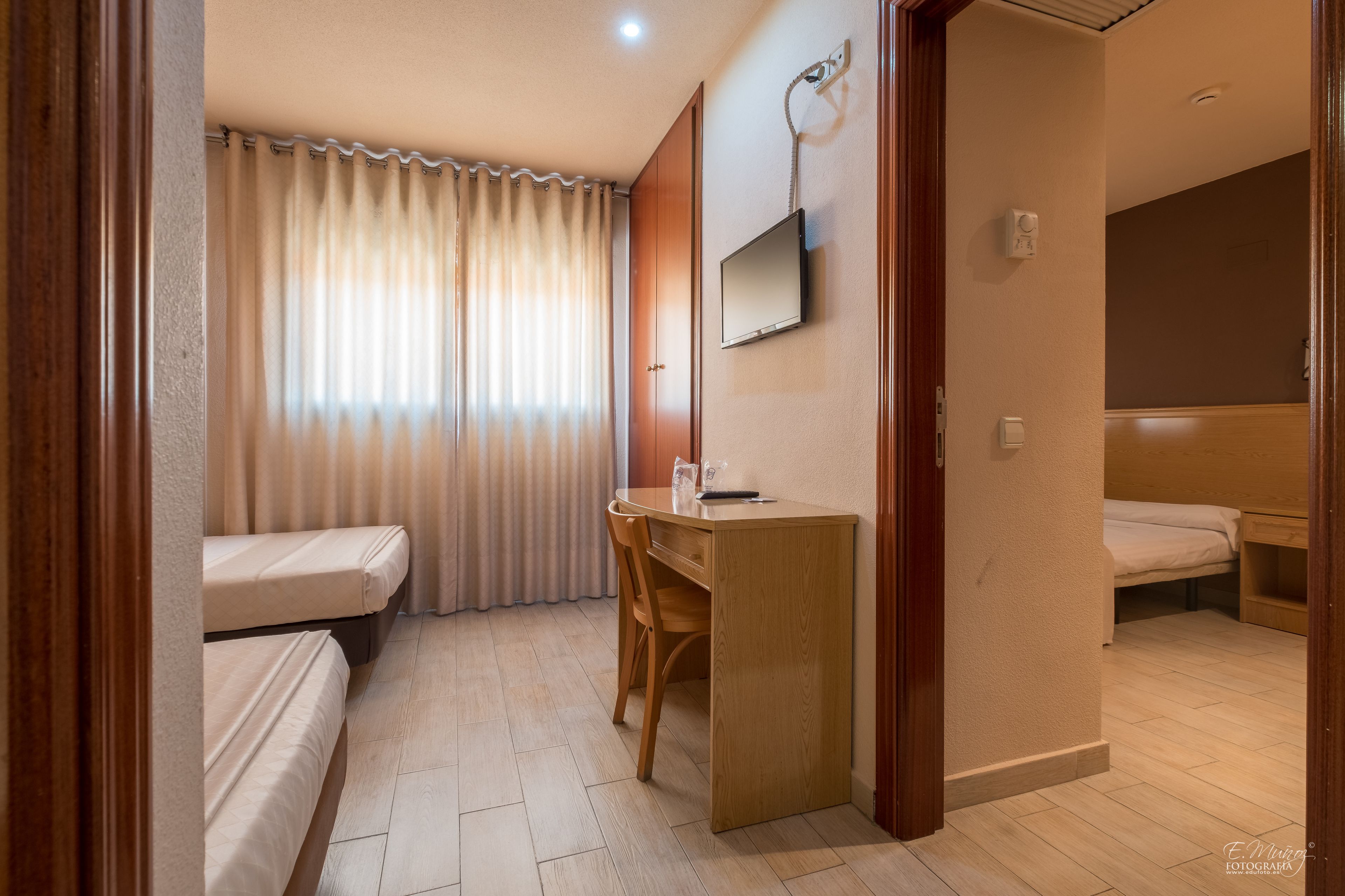 A hotel room with a wooden desk, a single bed, a second bed, a TV mounted on the wall, and a window with curtains.