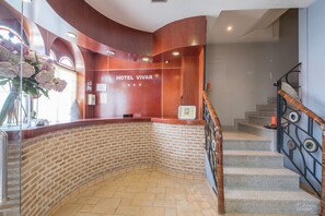 A hotel reception area with a brick wall, a wooden reception desk, and a staircase with a metal railing. - Hotel Vivar (Grinon)