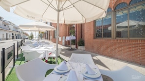 An outdoor dining area with white tables and umbrellas, a brick building, and a view of the street. - Hotel Vivar (Grinon)