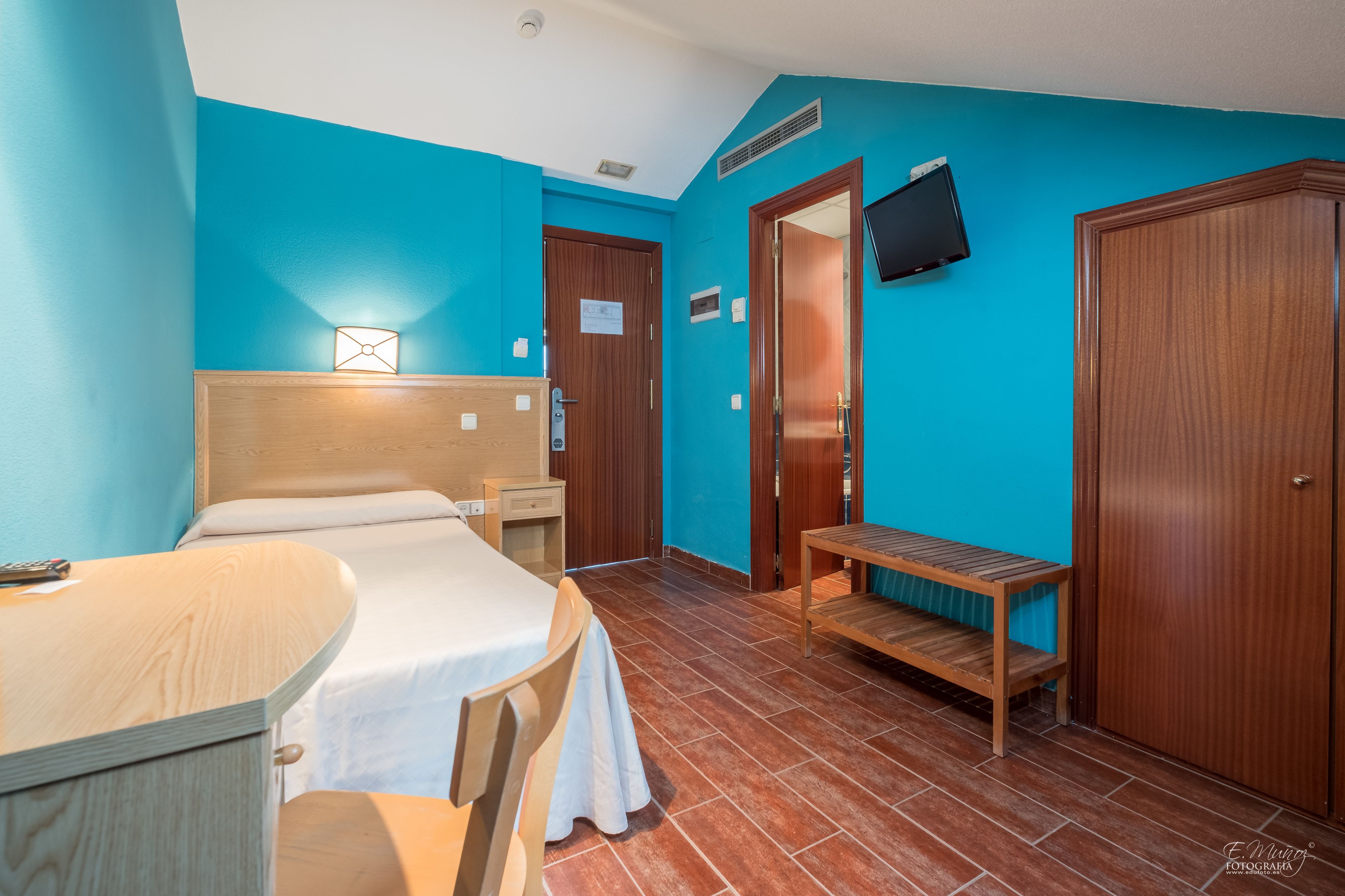 A hotel room with a bed, bedside table, chair, wooden bench, and a TV mounted on the wall.