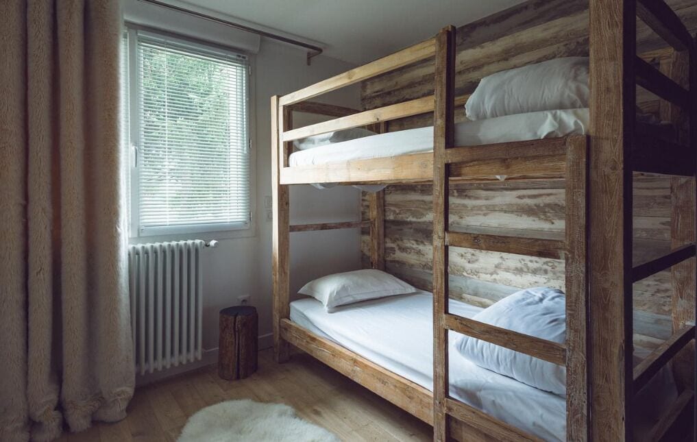 A bunk bed room with a rustic wooden design, a window with blinds, and a radiator.