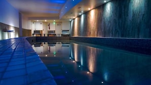 A modern indoor pool area with a reflective floor, seating area, and a wall with a textured finish. - Rock Noir Hôtel & Spa (La Salle-les-Alpes)