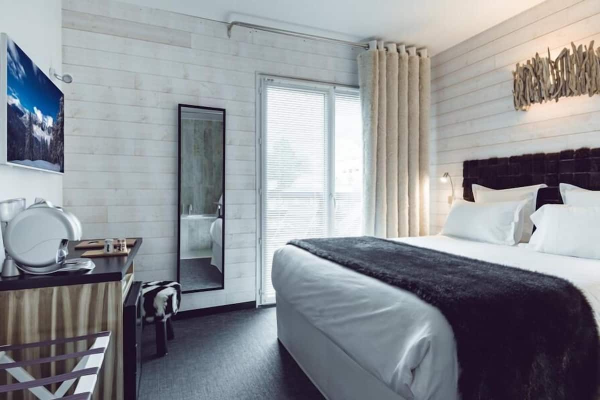 A modern hotel room with a large bed, a desk with a mirror, a wardrobe, and a window with curtains.