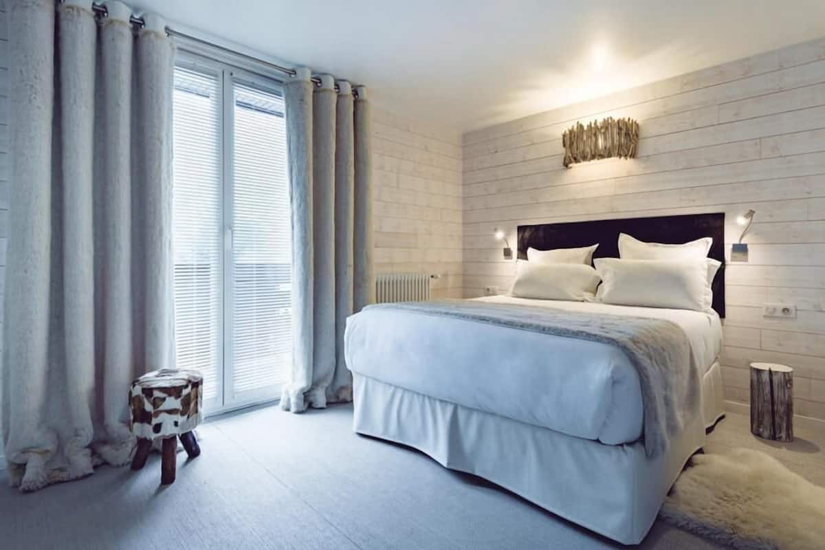A modern bedroom with a large bed, light-colored curtains, a small stool, and a rustic wall decor piece.