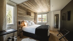 A bedroom with a wooden ceiling, a bed with white bedding, a black chair, a small table, and a fluffy rug. - Rock Noir Hôtel & Spa (La Salle-les-Alpes)