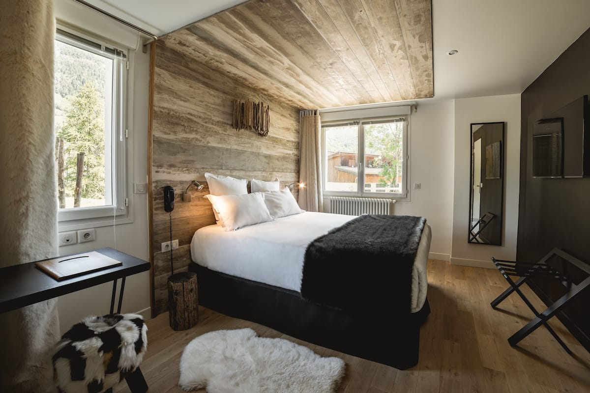A bedroom with a wooden ceiling, a bed with white bedding, a black chair, a small table, and a fluffy rug.