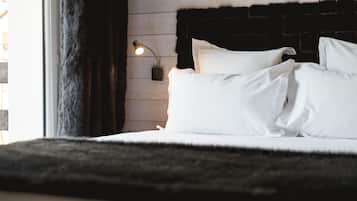 A neatly made bed with white linens and pillows, a black furry rug, a wall-mounted lamp, and a window with curtains.