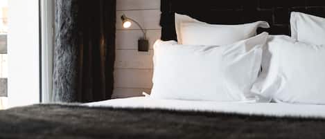 A neatly made bed with white linens and pillows, a black furry rug, a wall-mounted lamp, and a window with curtains.