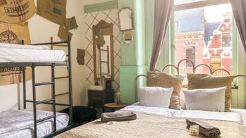 A dormitory room with bunk beds, a small table with a lamp, a mirror, and a window with a view of buildings.