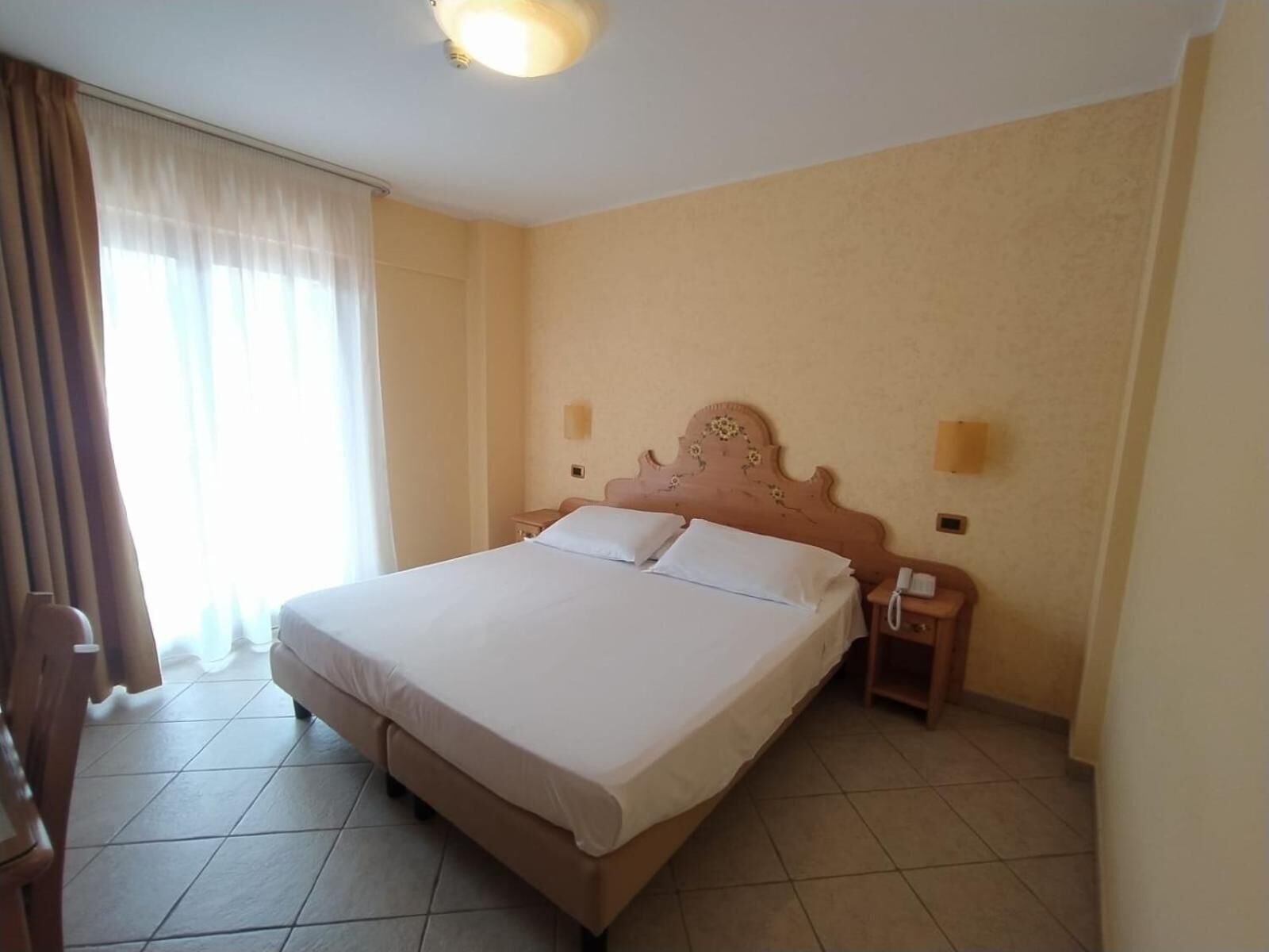 A hotel room with a large bed, two bedside tables, a nightstand, a wall-mounted telephone, and a window with curtains.