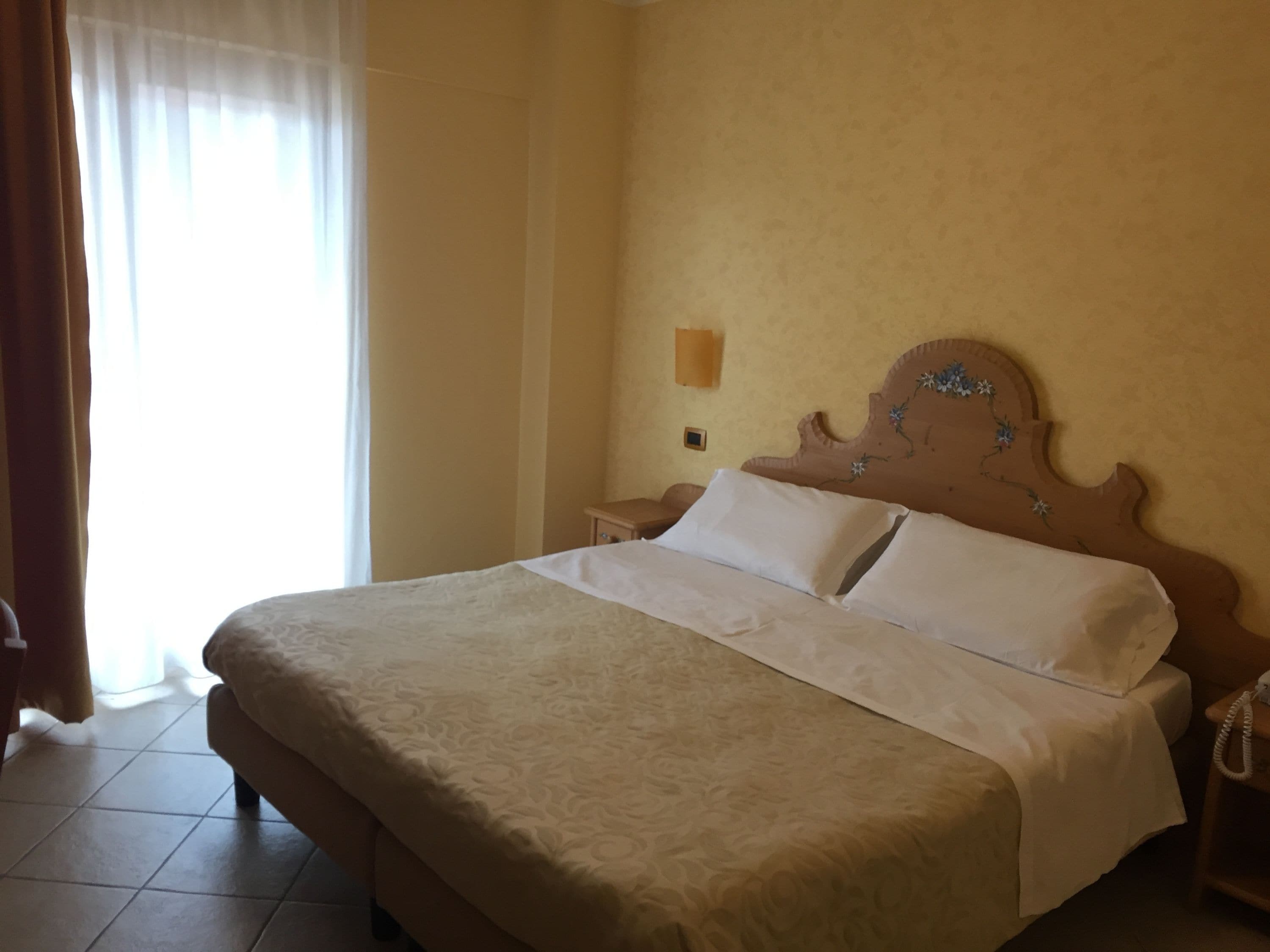 Camera d'albergo con un letto grande, un comodino, una lampada a parete e una finestra con tende.