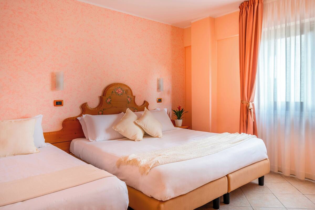 Une chambre d’hôtel avec deux lits, une tête de lit en bois, une plante en pot et une vue sur l’extérieur à travers des rideaux transparents.