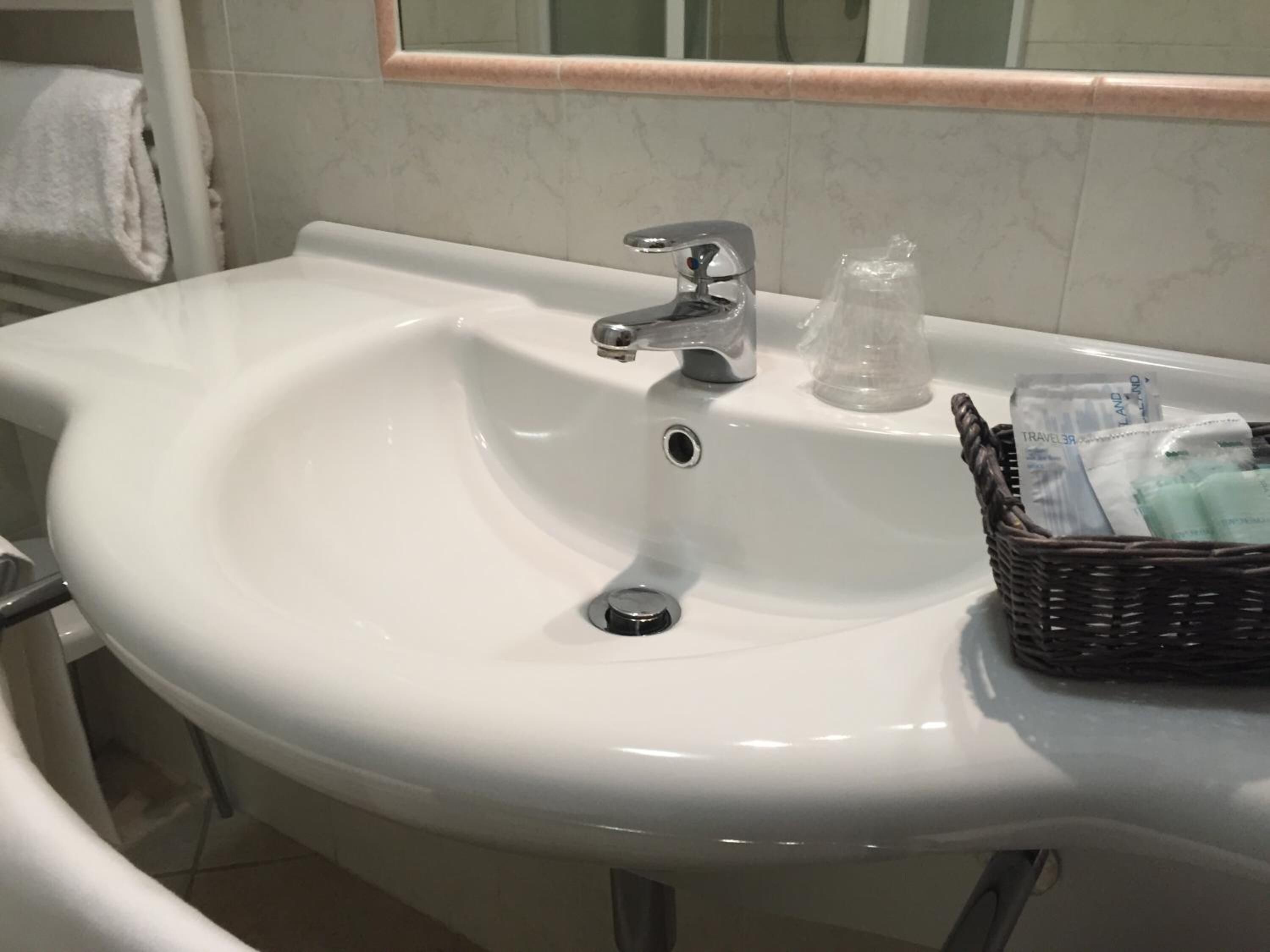 Uma pia branca com torneira cromada, um pequeno recipiente de vidro e uma cesta de vime com produtos de higiene pessoal.