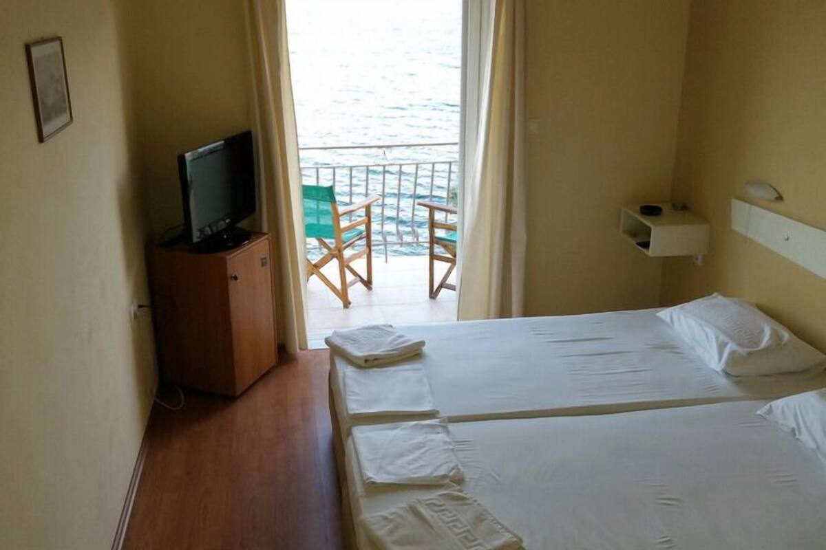 Habitación de hotel con dos camas, un televisor en un mueble, un balcón con una silla y una pequeña mesa.