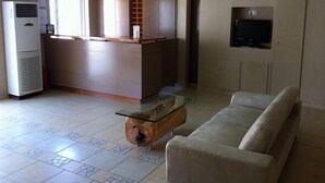 A modern living room with a glass coffee table, a beige sofa, a built-in TV, and a kitchen area with a refrigerator and a microwave. - Annema Hotel (Monemvasia)