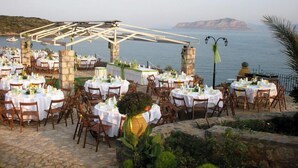 An outdoor dining area with round tables and chairs, set against a backdrop of a body of water and hills. - Annema Hotel (Monemvasia)