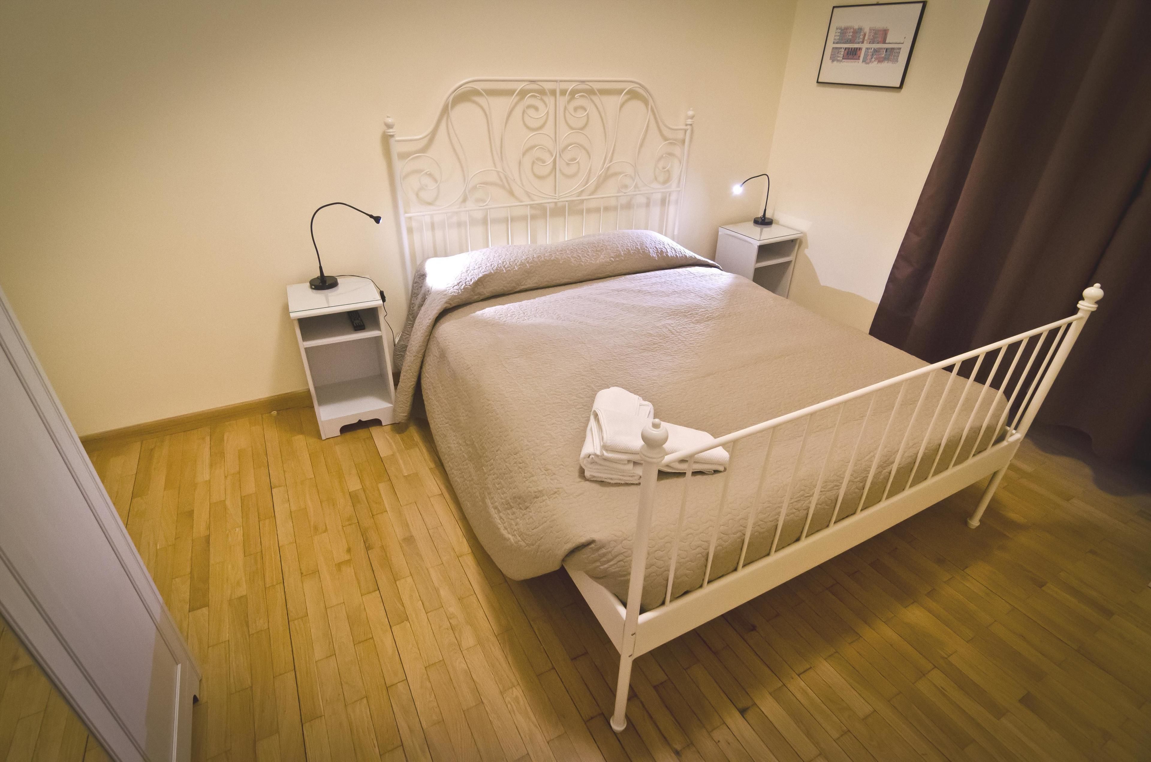 Una cama bien tendida con estructura metálica blanca y una manta color beige, dos mesitas de noche con lámparas, y suelo de madera.