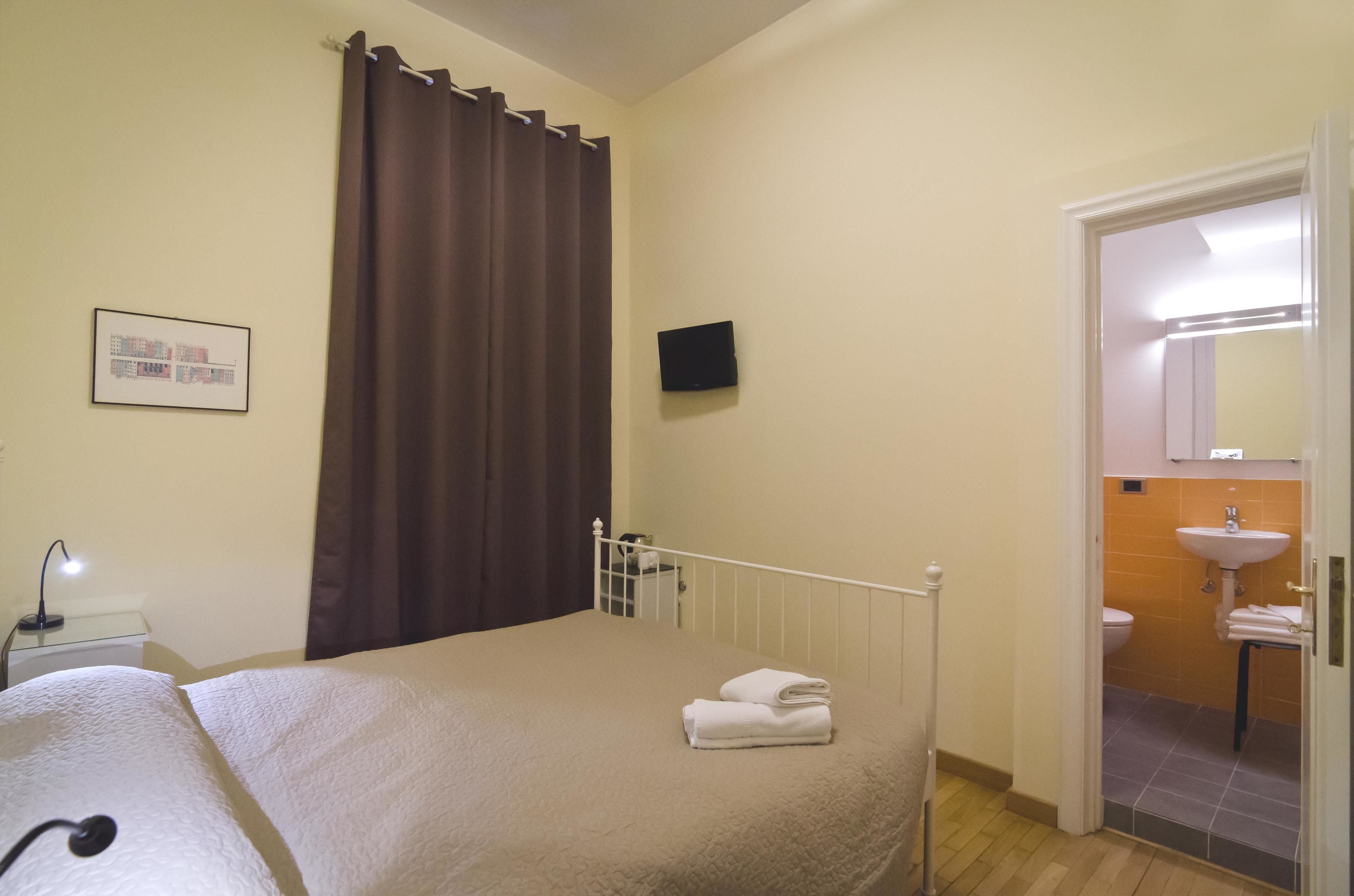 A hotel room with a bed, a bedside table with a lamp, a TV mounted on the wall, a bathroom with a sink and mirror, and a door leading to another room.