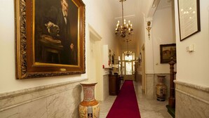 Un pasillo de mármol con alfombra roja, un cuadro grande enmarco y un jarrón decorativo.