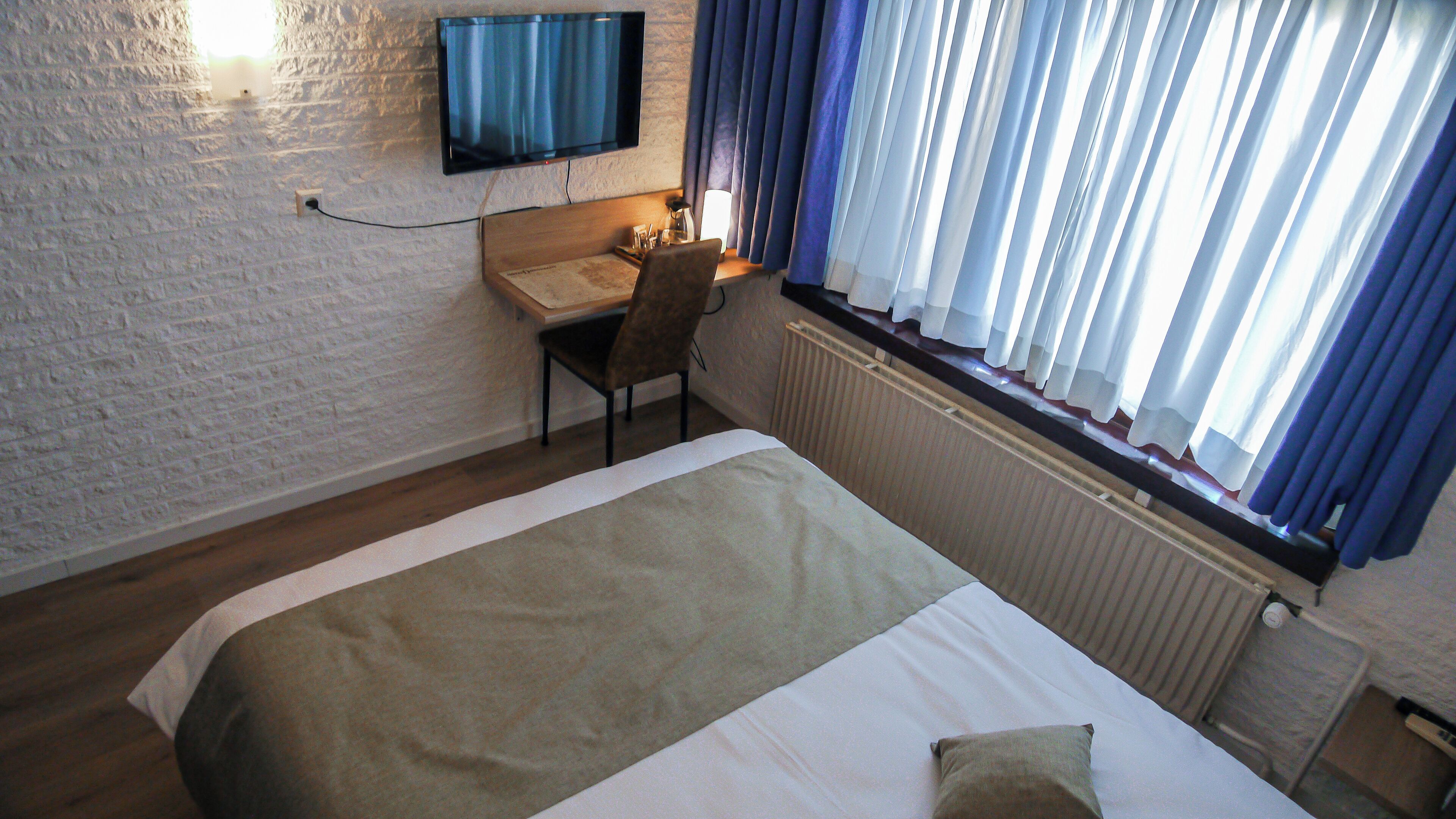 A hotel room with a bed, a desk, a chair, a TV mounted on the wall, and a window with curtains.