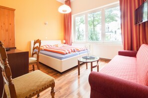A bedroom with a bed, a wooden wardrobe, a chair, a red sofa, a small table, and a window with curtains. - Hotel Marco Polo (Hamburg)