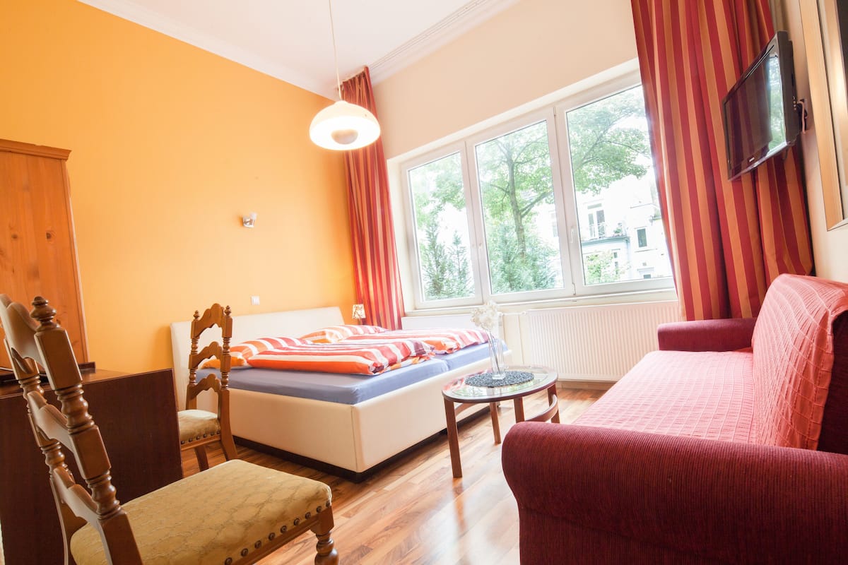 A room with a bed, a wooden chair, a red sofa, a small table, and a TV mounted on the wall.
