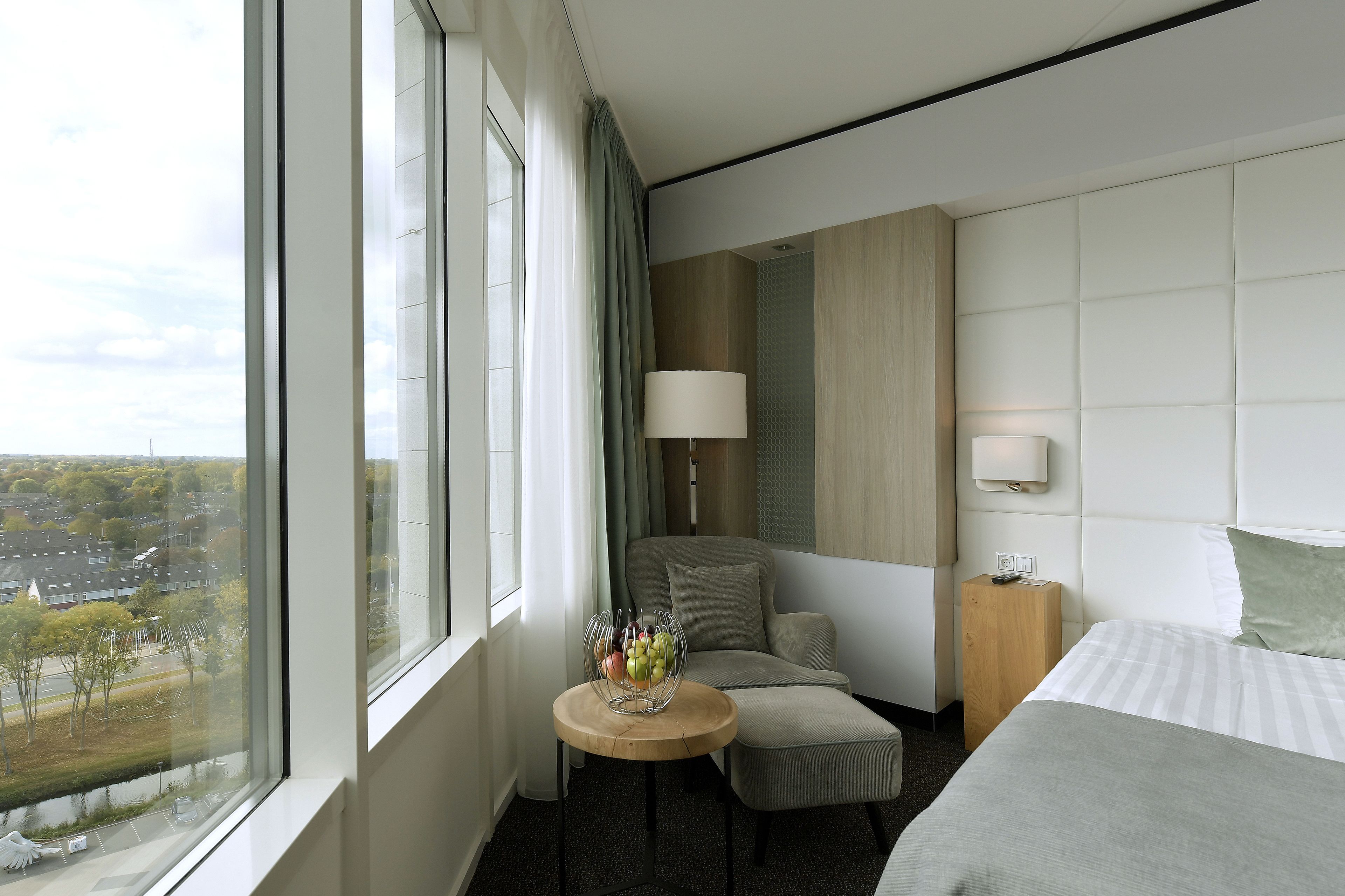 A hotel room with a large window, a bed, a sofa, a small table with fruit, and a lamp.