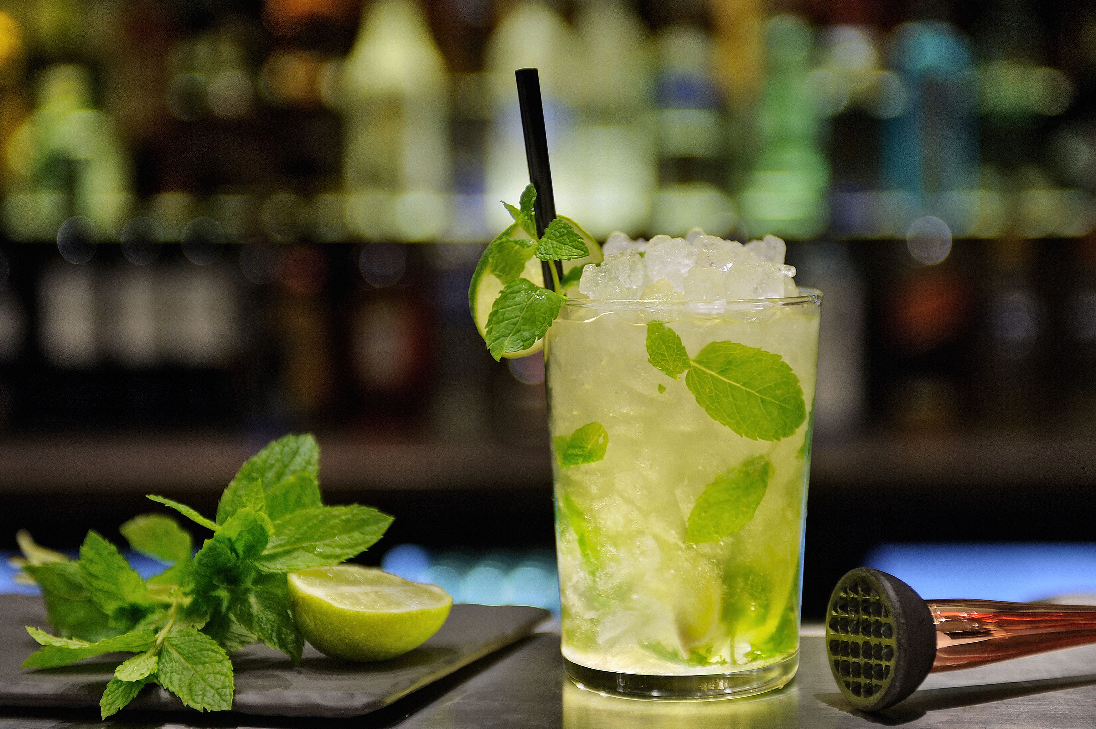 Een cocktail met muntblaadjes, ijsblokjes, een limoenpartje en een cocktailshaker op een bar.