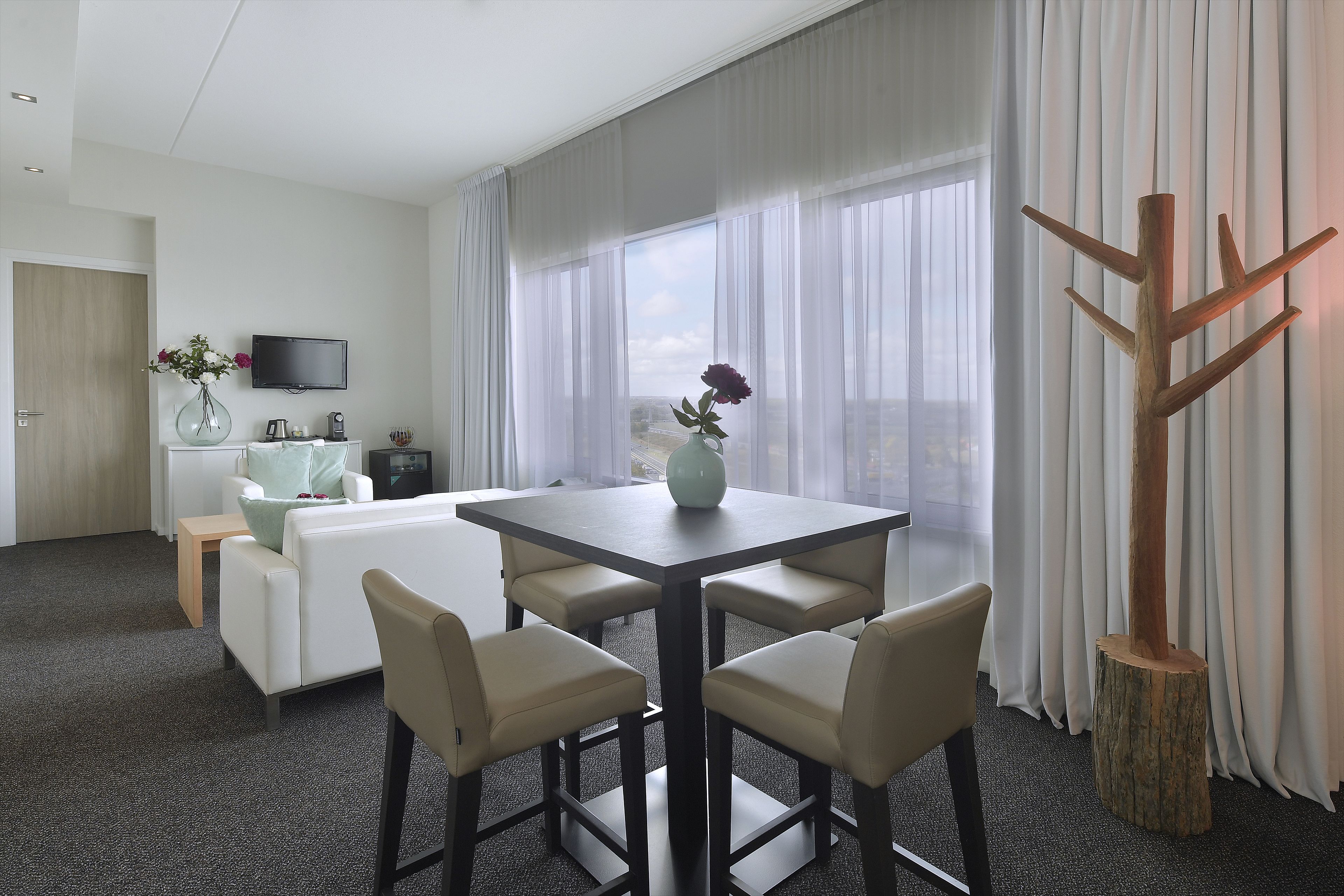 Une chambre d’hôtel moderne équipée d’une table à manger, de quatre chaises, d’un canapé, d’une télévision et d’une grande fenêtre avec des rideaux.