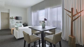 Une chambre d’hôtel moderne équipée d’une table à manger, de quatre chaises, d’un canapé, d’une télévision et d’une grande fenêtre avec des rideaux.