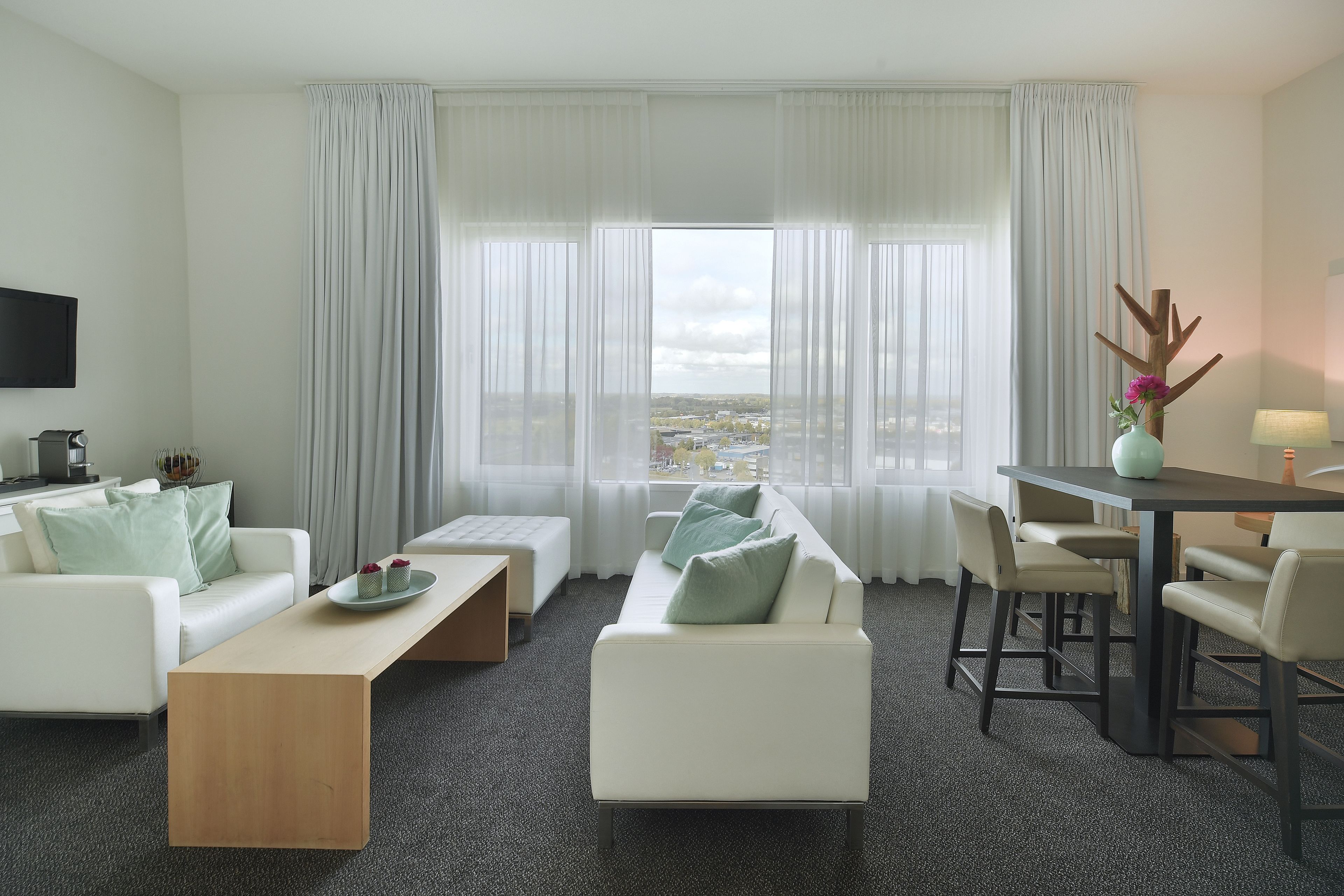 Een moderne hotelkamer met een witte bank, een houten bijzettafeltje, een eettafel met stoelen en een groot raam met dunne gordijnen.