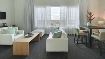 Een moderne hotelkamer met een witte bank, een houten bijzettafeltje, een eettafel met stoelen en een groot raam met dunne gordijnen.