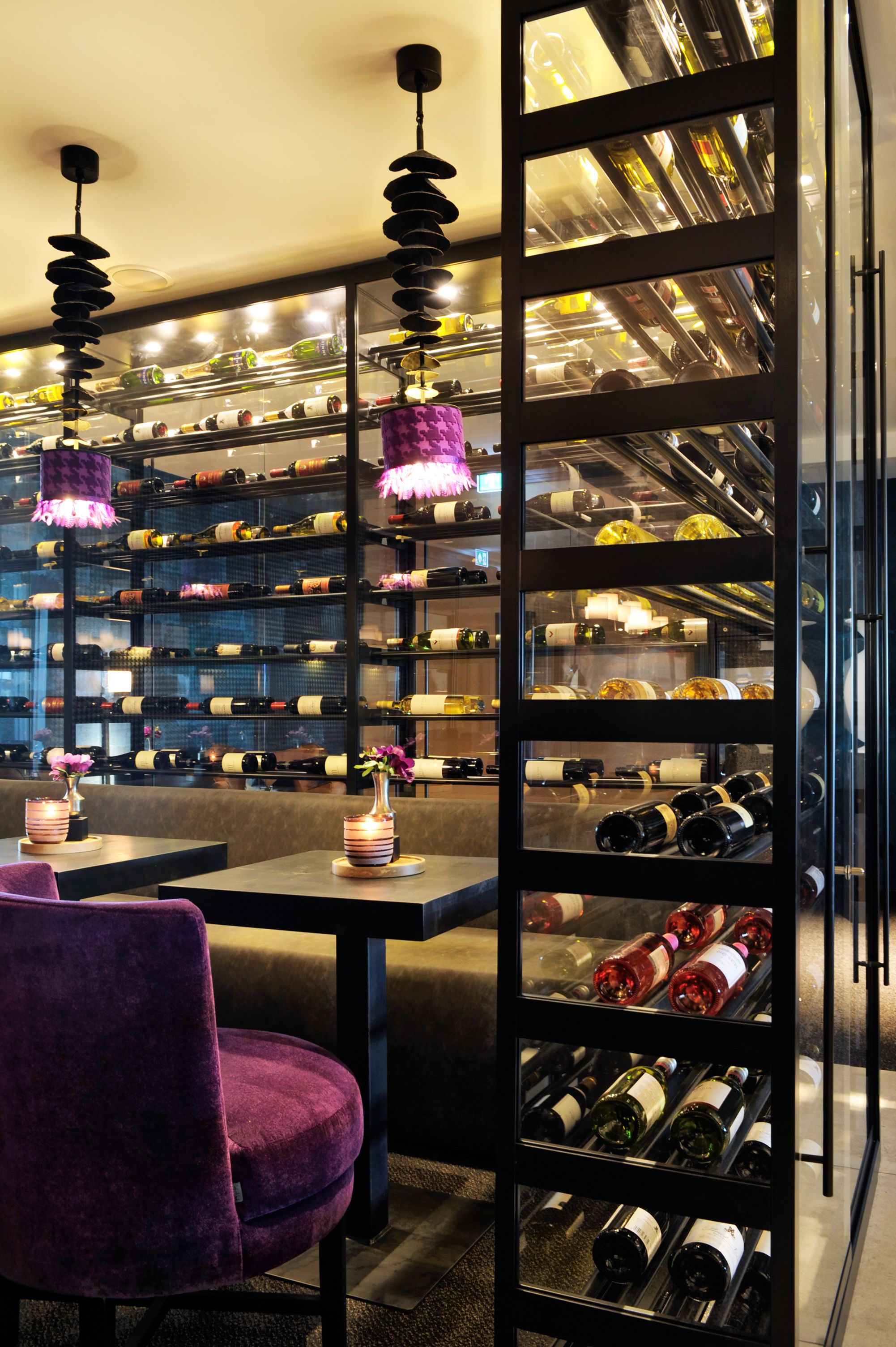 Une cave à vin avec des étagères remplies de bouteilles, un coin salon avec un fauteuil violet et des lampes suspendues.