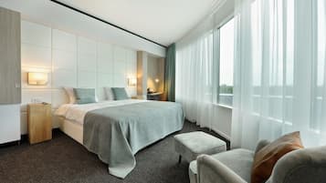 A modern hotel room with a large bed, a sitting area with a chair, a bedside table, and a view of the outdoors through sheer curtains.