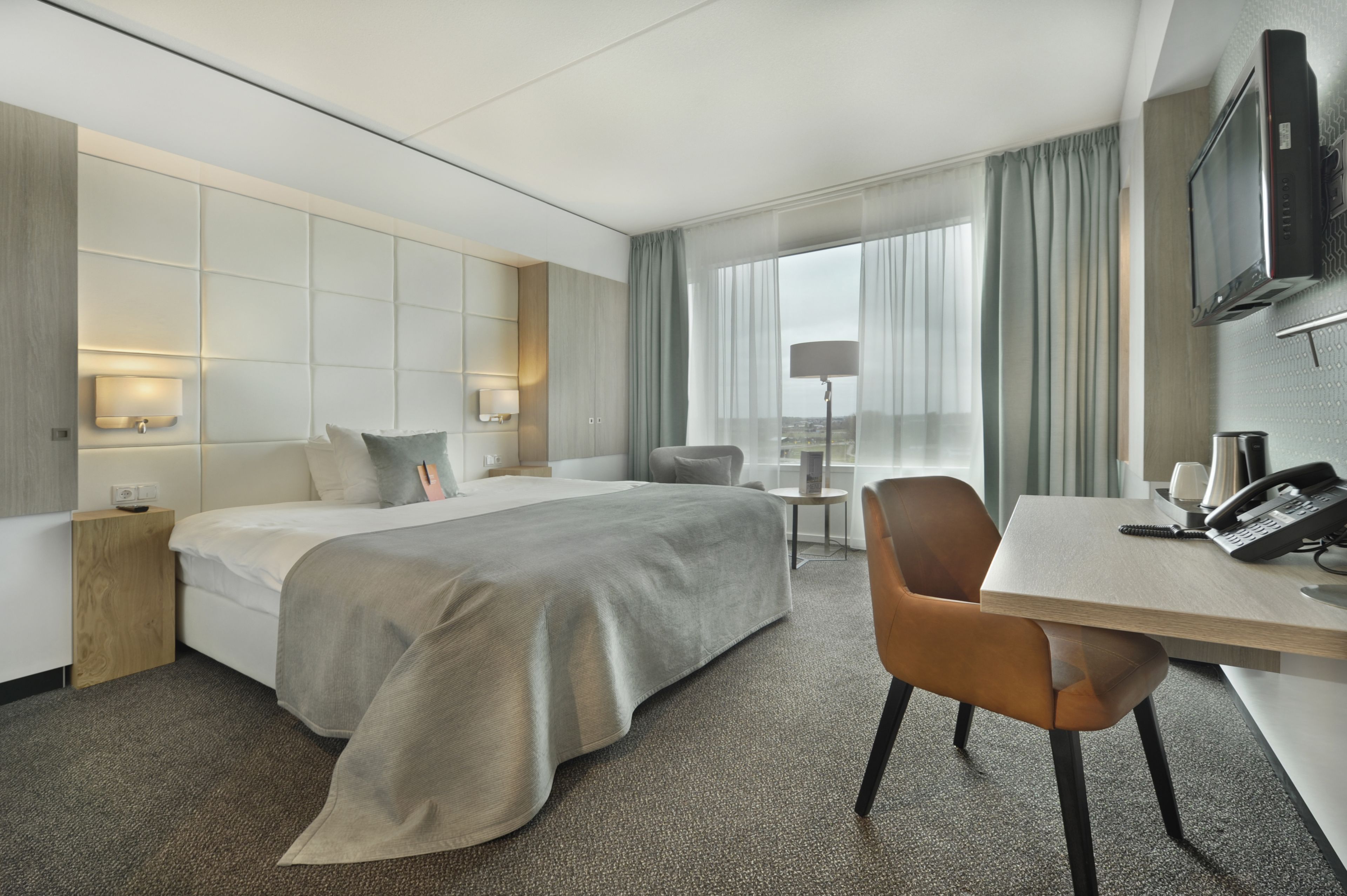 A modern hotel room with a large bed, a desk with a telephone, a chair, a small table, and a TV mounted on the wall.