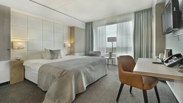A modern hotel room with a large bed, a desk with a telephone, a chair, a small table, and a TV mounted on the wall.