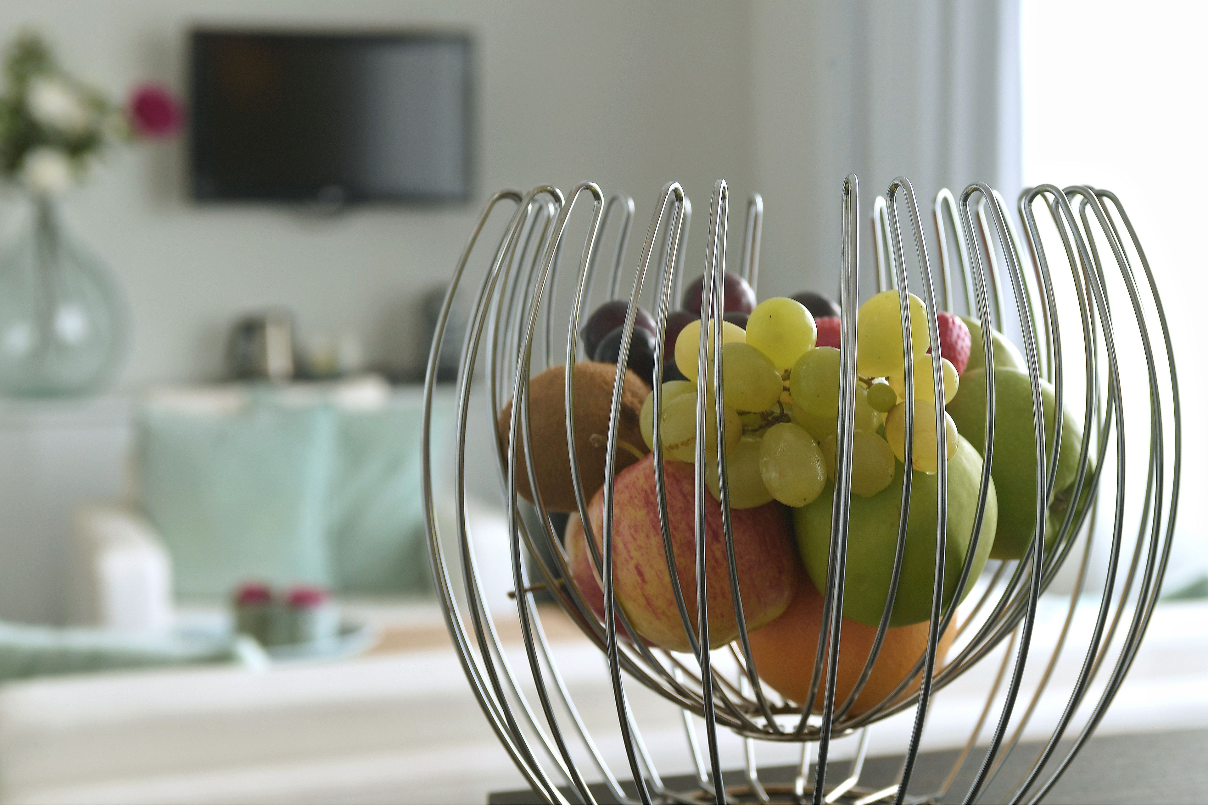 Een schaal vol druiven, appels en peren op een tafel in een kamer met een televisie en een vaas op de achtergrond.